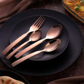 Elegant rose gold cutlery set displayed on black dinnerware, styled with gourmet Indian dishes.